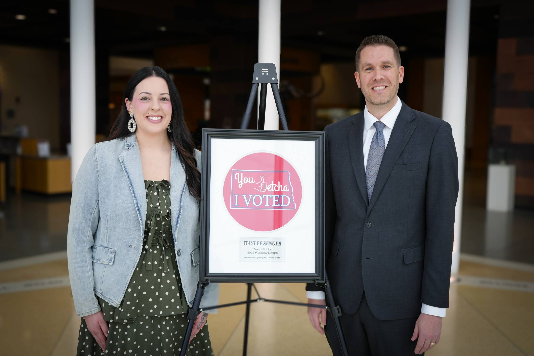 Haylee Senger and Secretary of State Michael Howe standing next to a framed image of Haylee's winning sticker design.