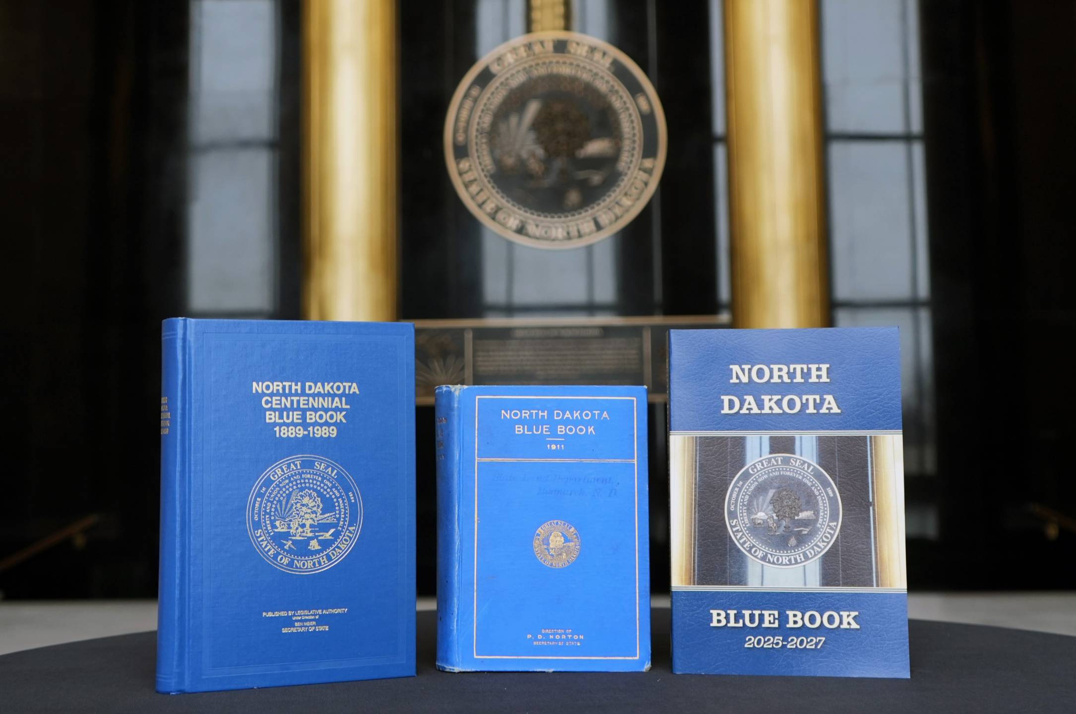 Three versions of the North Dakota Blue Book. The Centennial edition is blue with a hard cover and an embossed gold North Dakota Great Seal. The 1911 edition of the Blue Book features the original embossed Great Seal. The 2025-2027 North Dakota Blue Book has a photograph of the bronze Great Seal that hangs in the State Capitol Memorial Hall.
