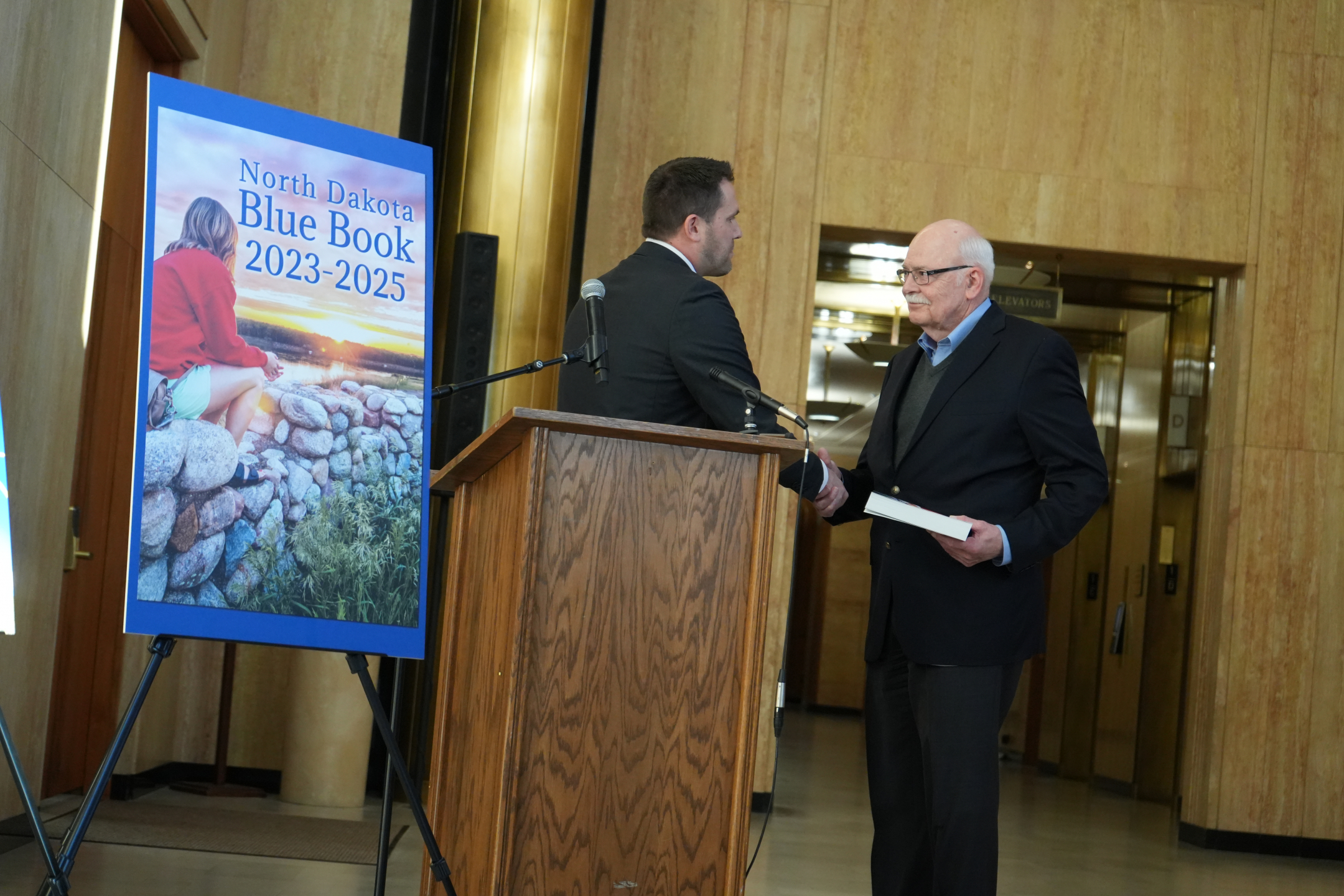 Secretary of State Michael Howe is shaking the hand of former Secretary of State Al Jaeger, who is holding a copy of the blue book. Both men are dressed in business suits.