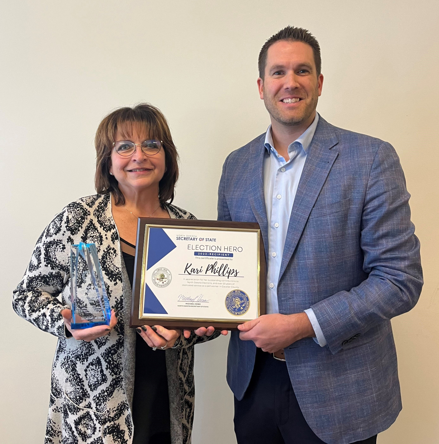 Kari Phillips, a female, is standing on the left. Secretary of State Michael Howe is standing on the right. They are holding a certificate between them and Kari is also holding a glass award in her other hand.